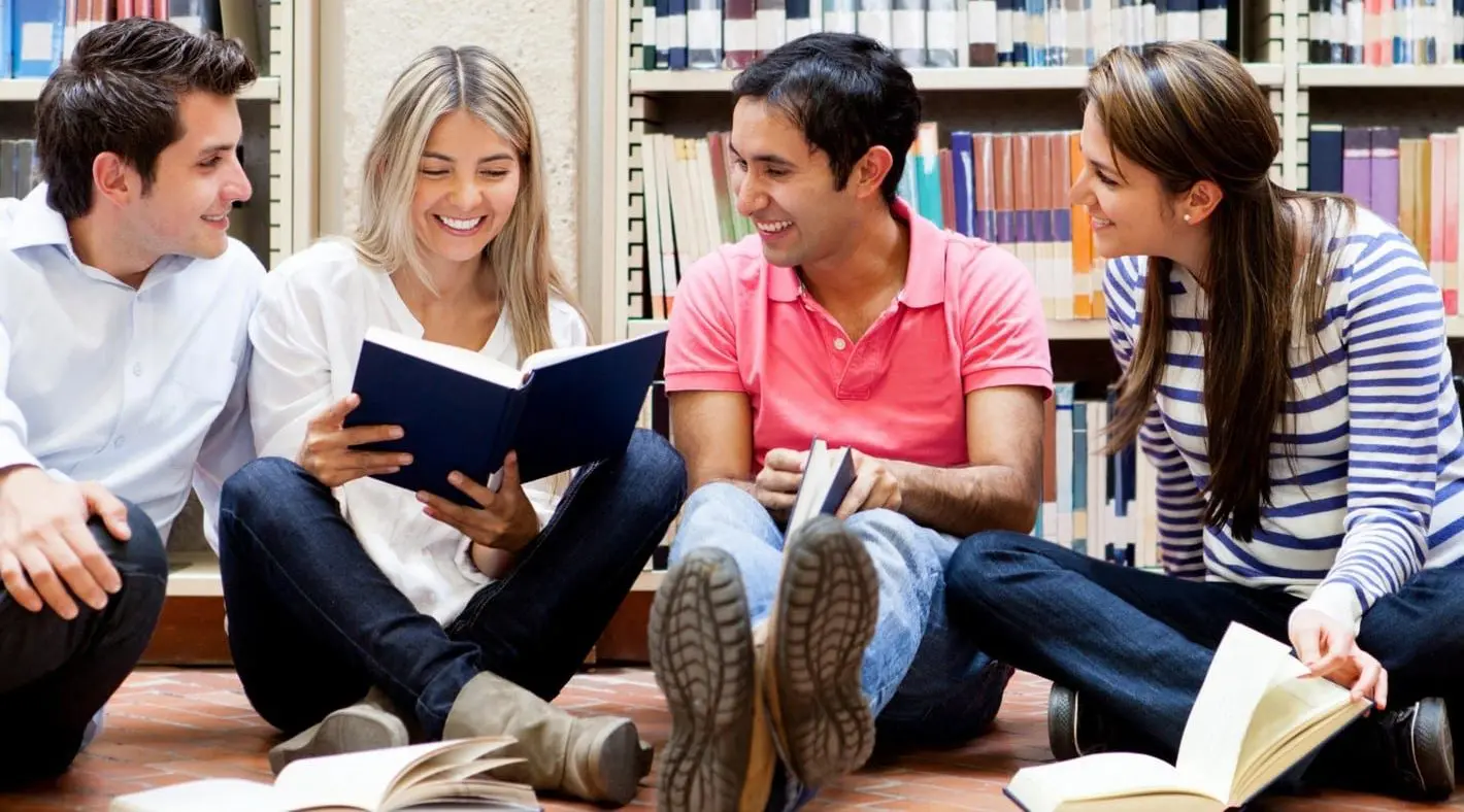 Students sat in a library, reading a book and smiling.